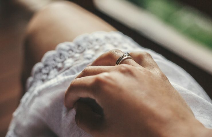 Foto de uma mão com anel de noivado no vestido boêmio branco de uma mulher na varanda de de madeira, nas montanhas e no verão. Anel de prata com cristal.